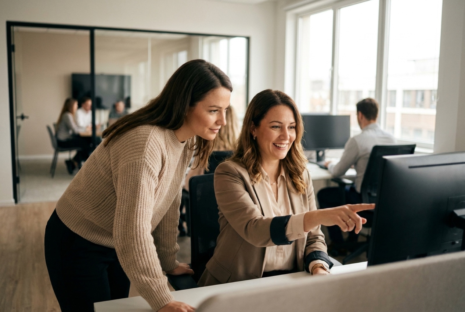 Two HR business partners collaborating over a large monitor in a glass-walled meeting room, one gesturing while they compare notes, warm afternoon daylight
