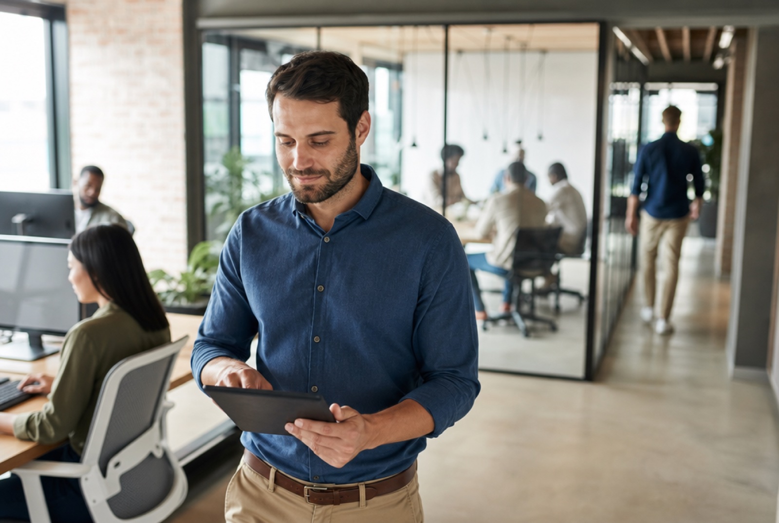 A team lead and an L&D specialist planning together at a shared desk, one clicking a laptop trackpad while the other holds a coffee, candid corporate-documentary style in a bright office