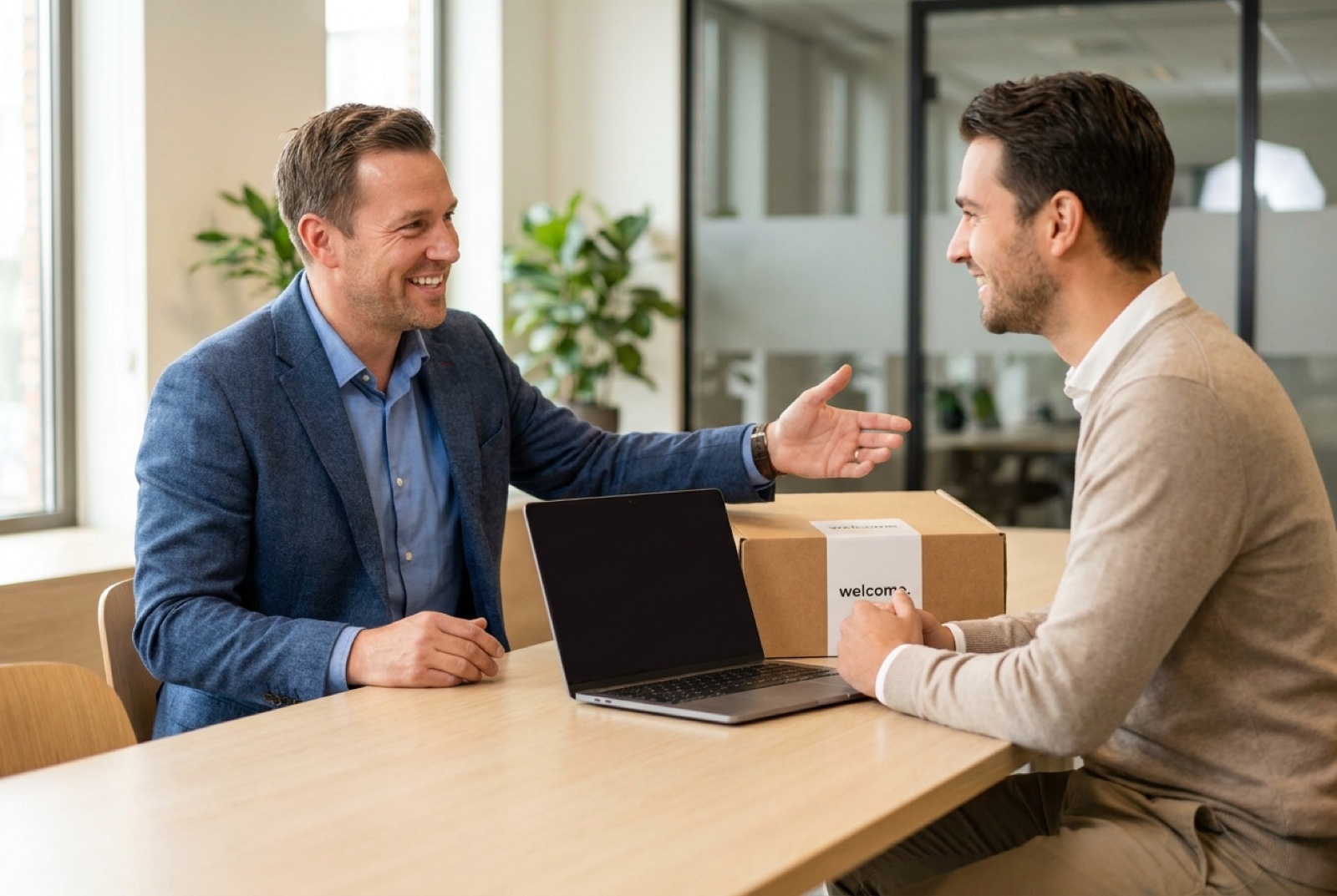 Manager warmly welcoming a new hire at a bright desk on their first morning, a welcome kit and laptop on the desk, open-plan office softly blurred behind