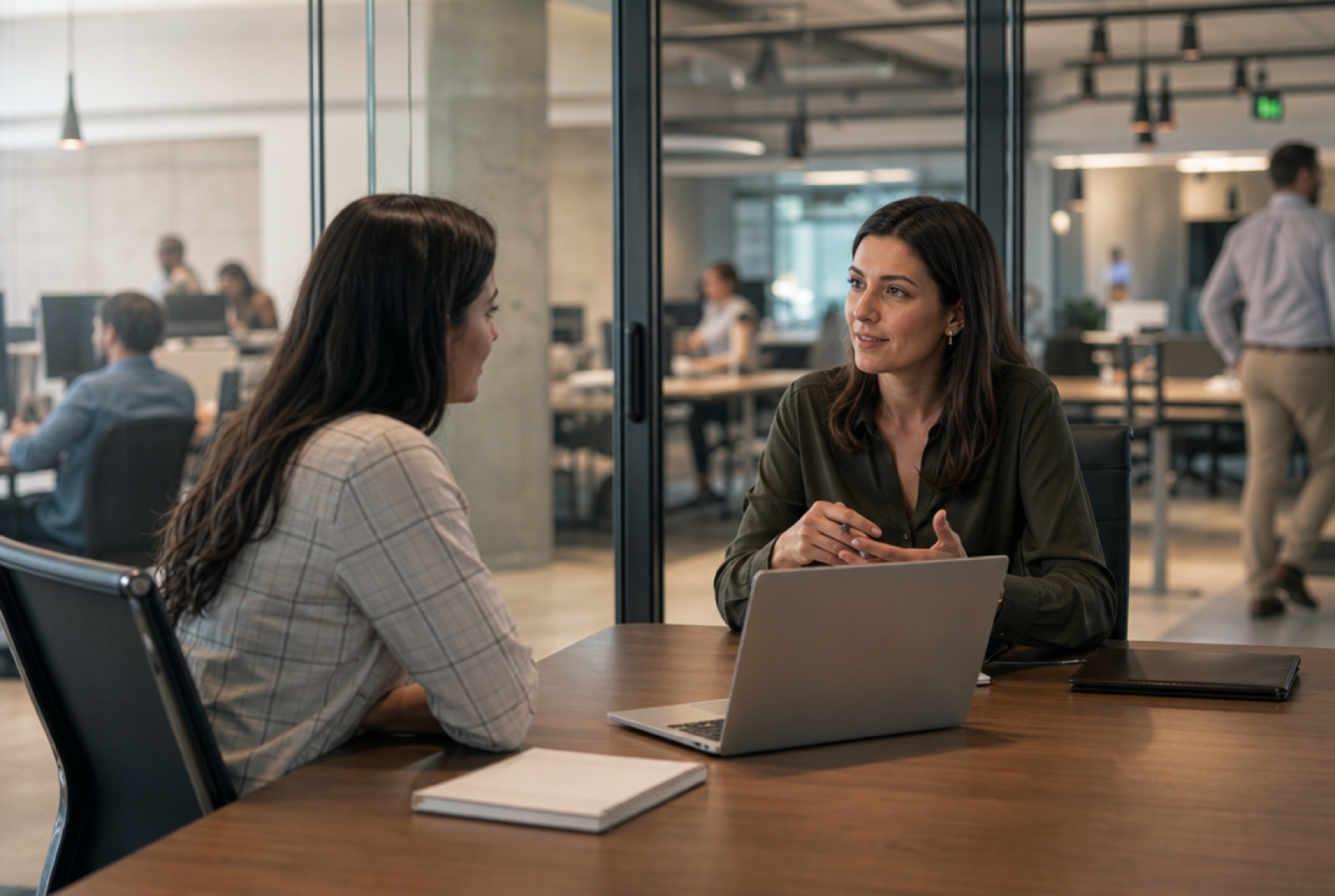 A sales manager and a team member in a focused one-on-one at a desk, laptop open between them, both leaning in, open-plan office softly blurred behind