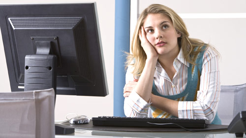 Bored young woman at office desk, looking away from computer with a thoughtful expression.