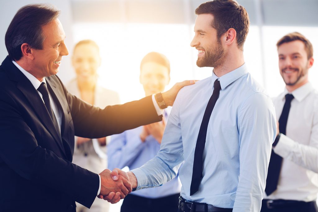 Two businessmen shaking hands with colleagues smiling in the background.