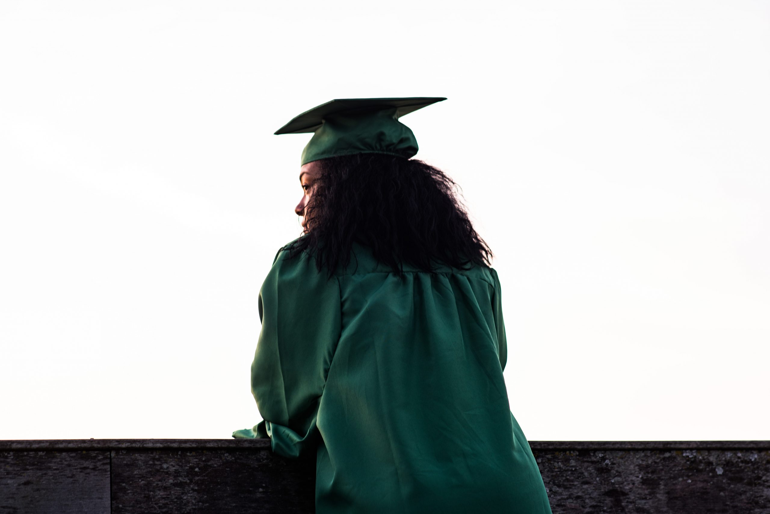 Graduate in green gown looking out contemplatively, symbolizing future aspirations.