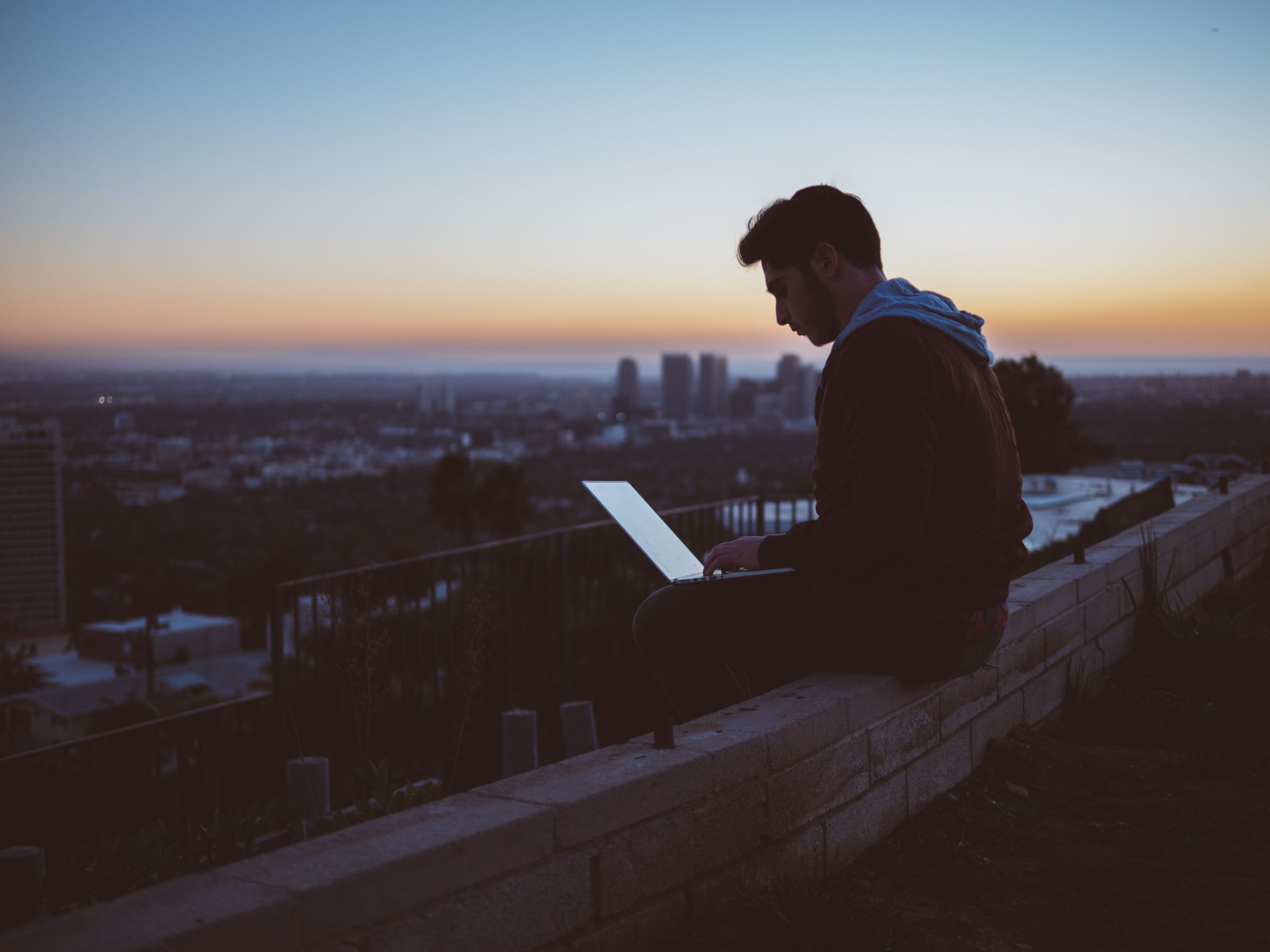 Man working on laptop at sunset with cityscape in background.