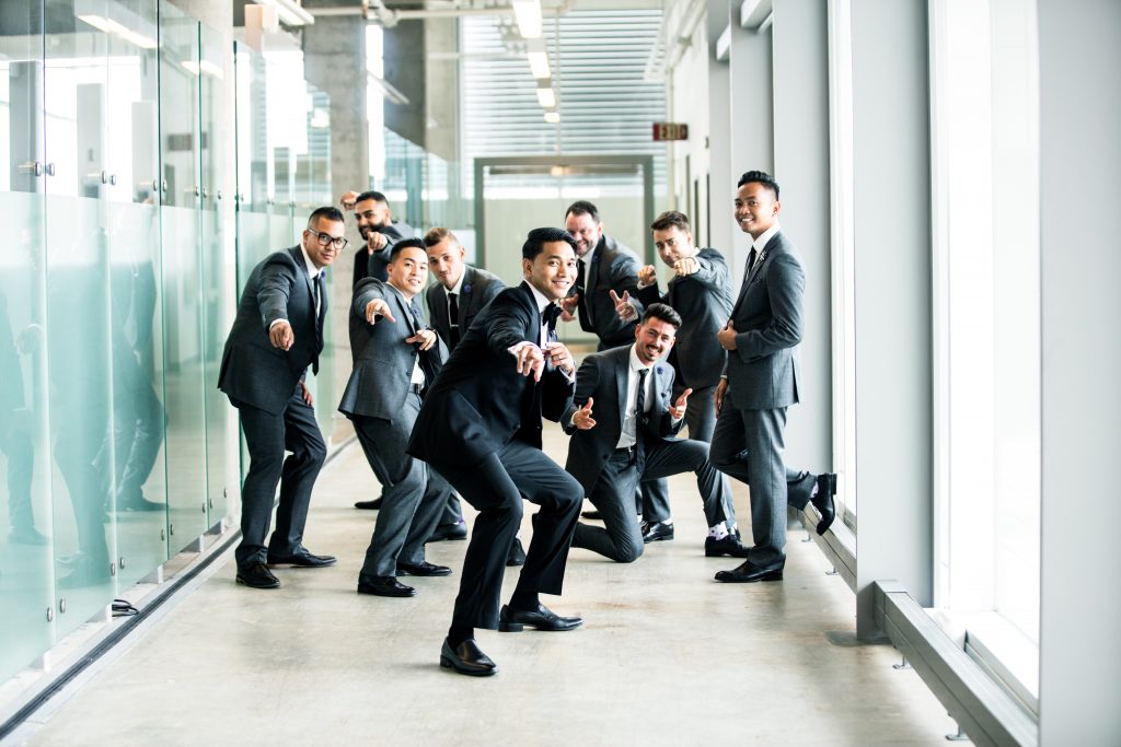 Group of cheerful businessmen in suits posing playfully in a modern office corridor.