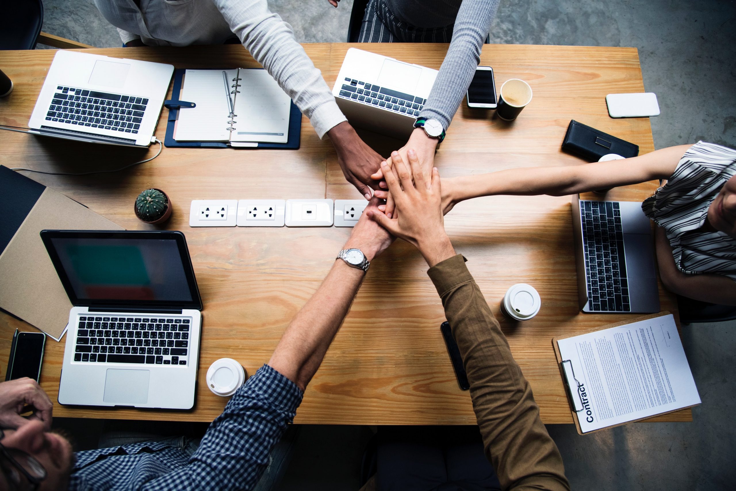 Team placing hands together over a table with laptops and work documents.