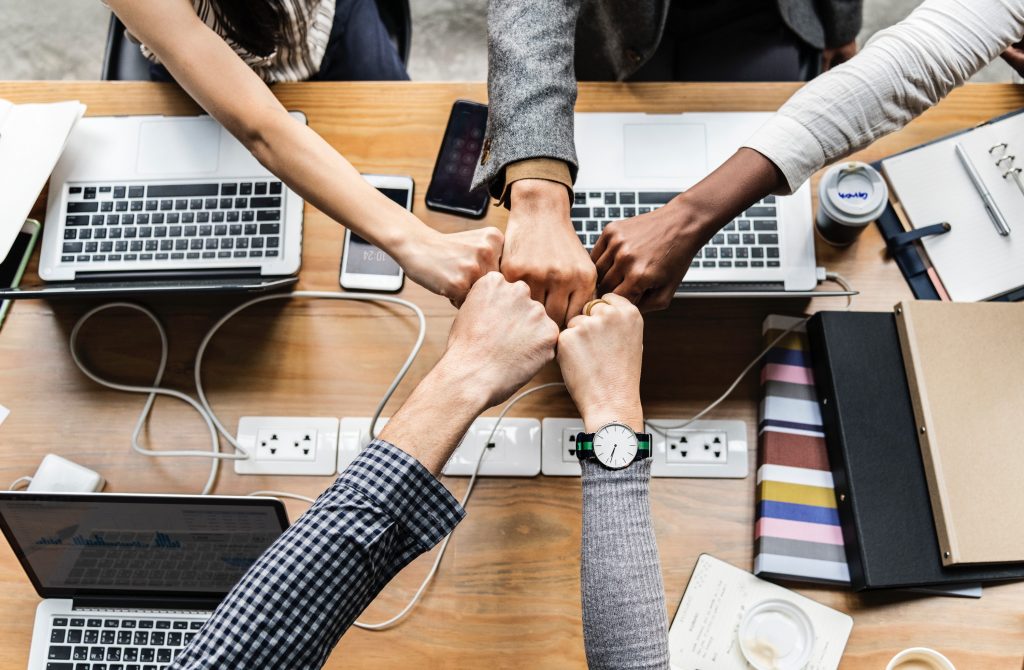 Diverse team fists together over workspace with laptops and notebooks.