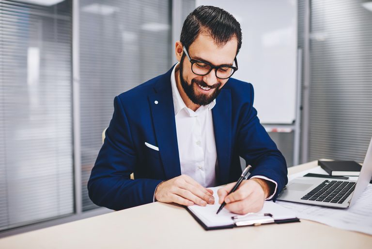 Smiling businessman writing notes with laptop on desk.