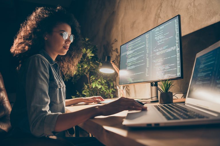 Focused woman programming on multiple computer screens in a dark room.