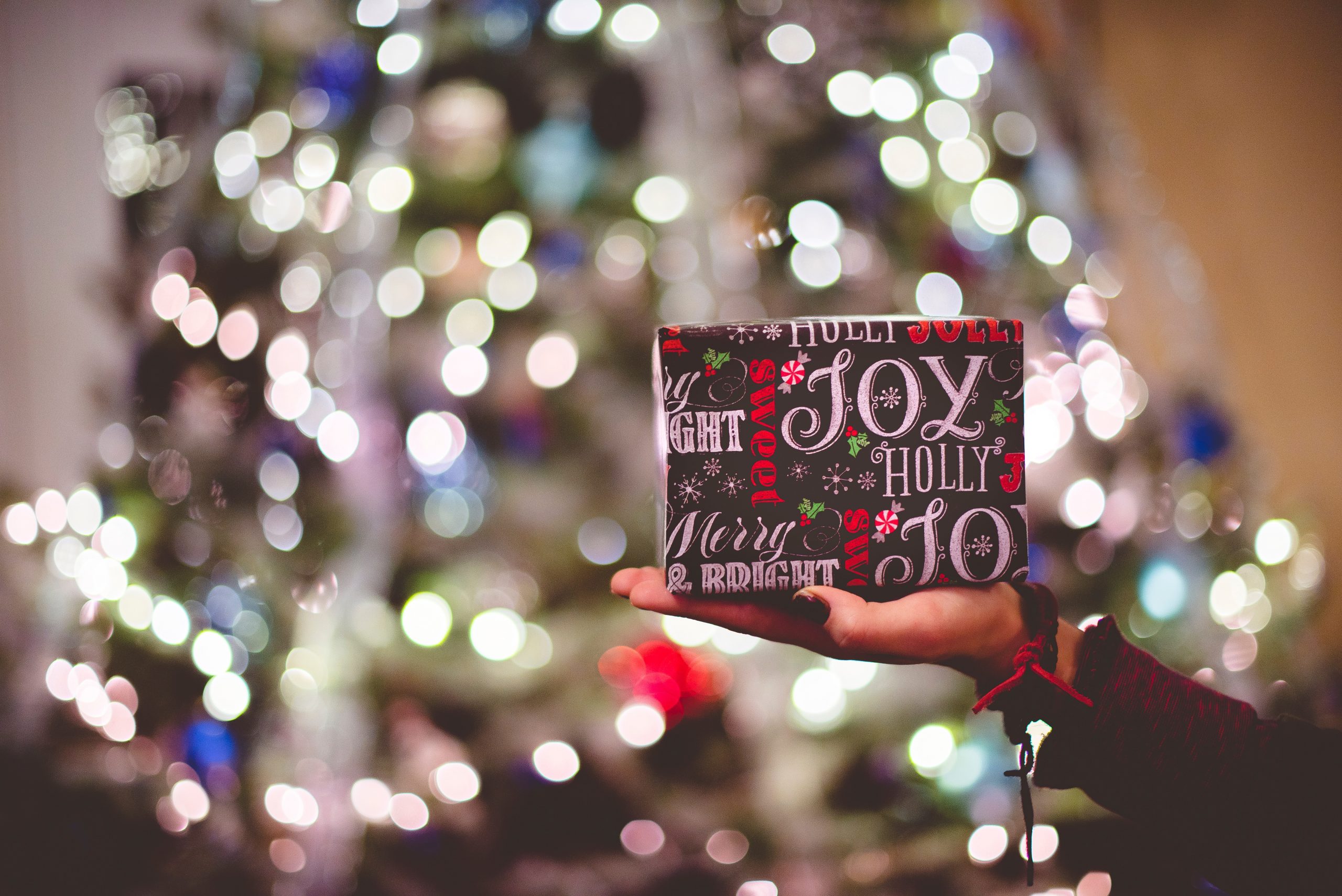 Hand holding a festive gift box with 'Joy, Holly, Jolly' text against a blurry Christmas tree backdrop.