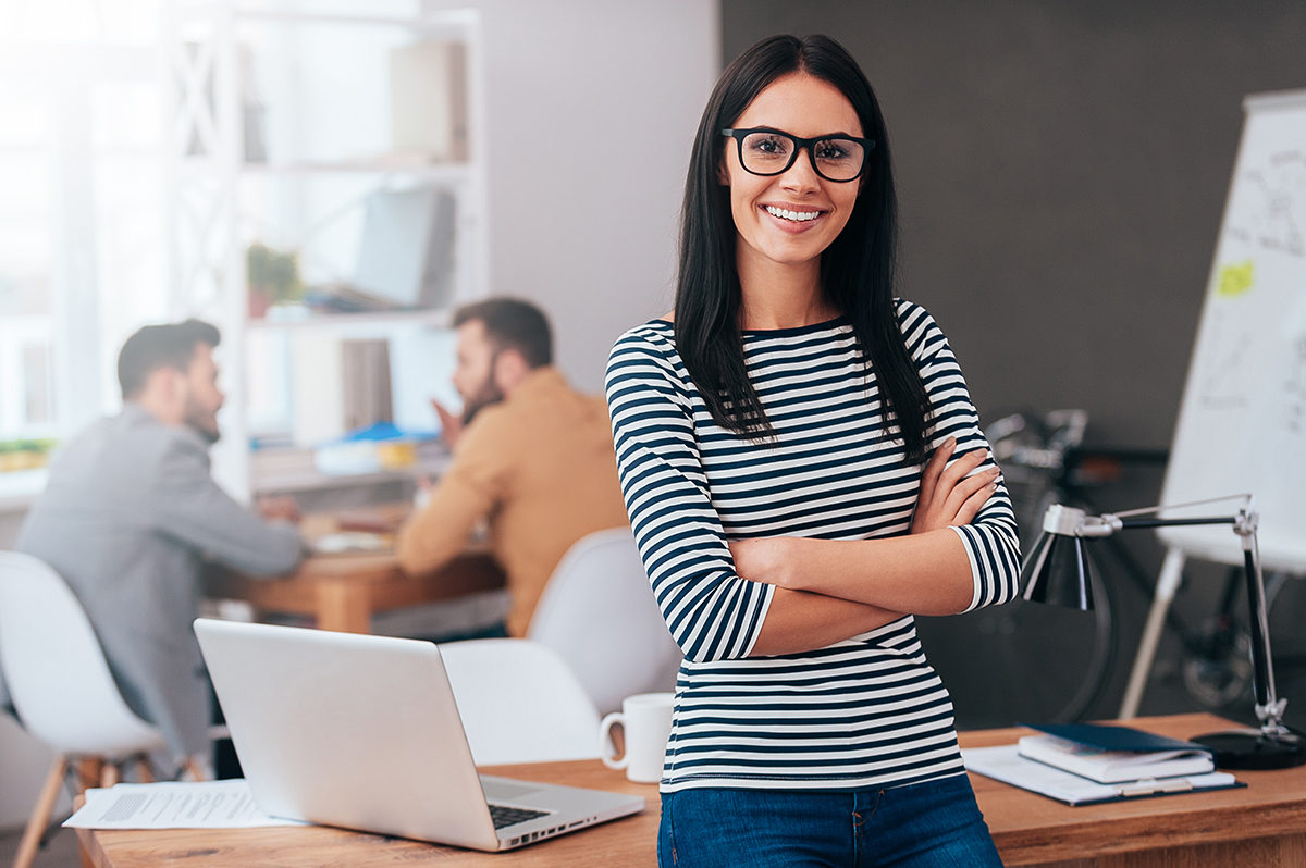 Confident woman in striped top with crossed arms, office meeting in background.