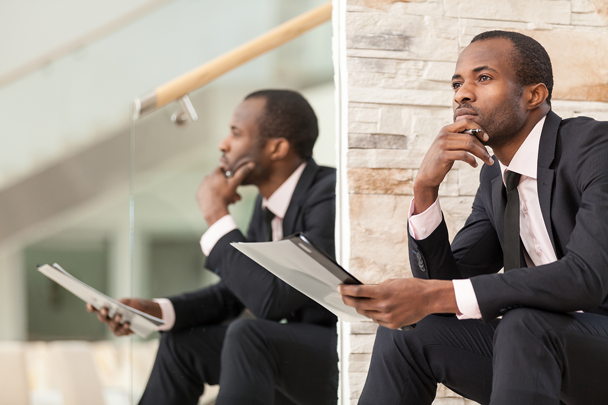 Contemplative businessman holding a tablet while sitting against a reflective surface.