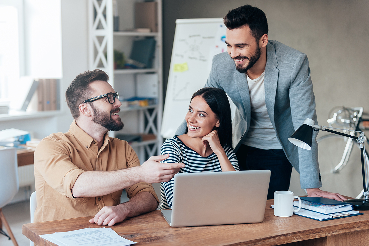 Three colleagues with a laptop smiling and discussing in a bright office.