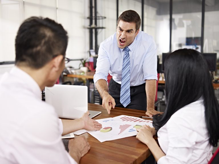 Angry manager yelling and pointing at colleagues during a tense meeting with charts on the table.
