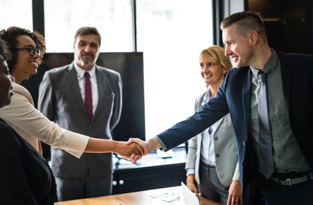 Two professionals shaking hands in a meeting with colleagues.