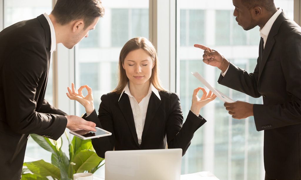 Businesswoman meditating at work with colleagues showing 'okay' sign.