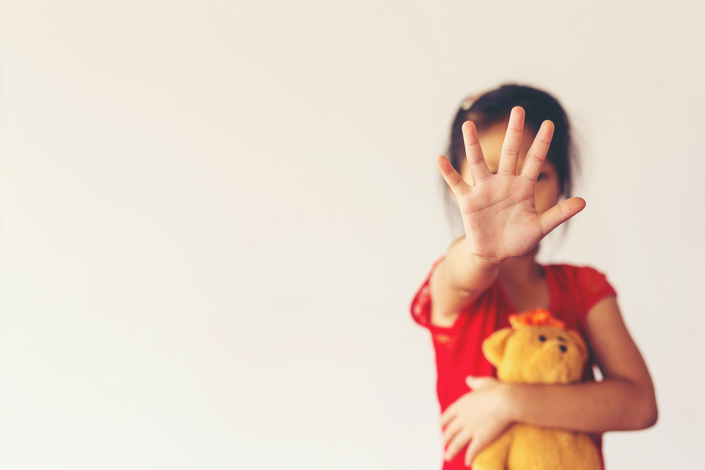 Child holding up hand in stop gesture with blurred teddy bear.