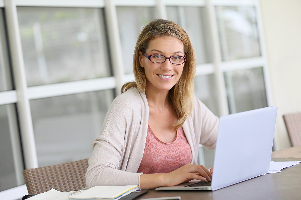 Smiling woman with glasses working on laptop at bright office desk.