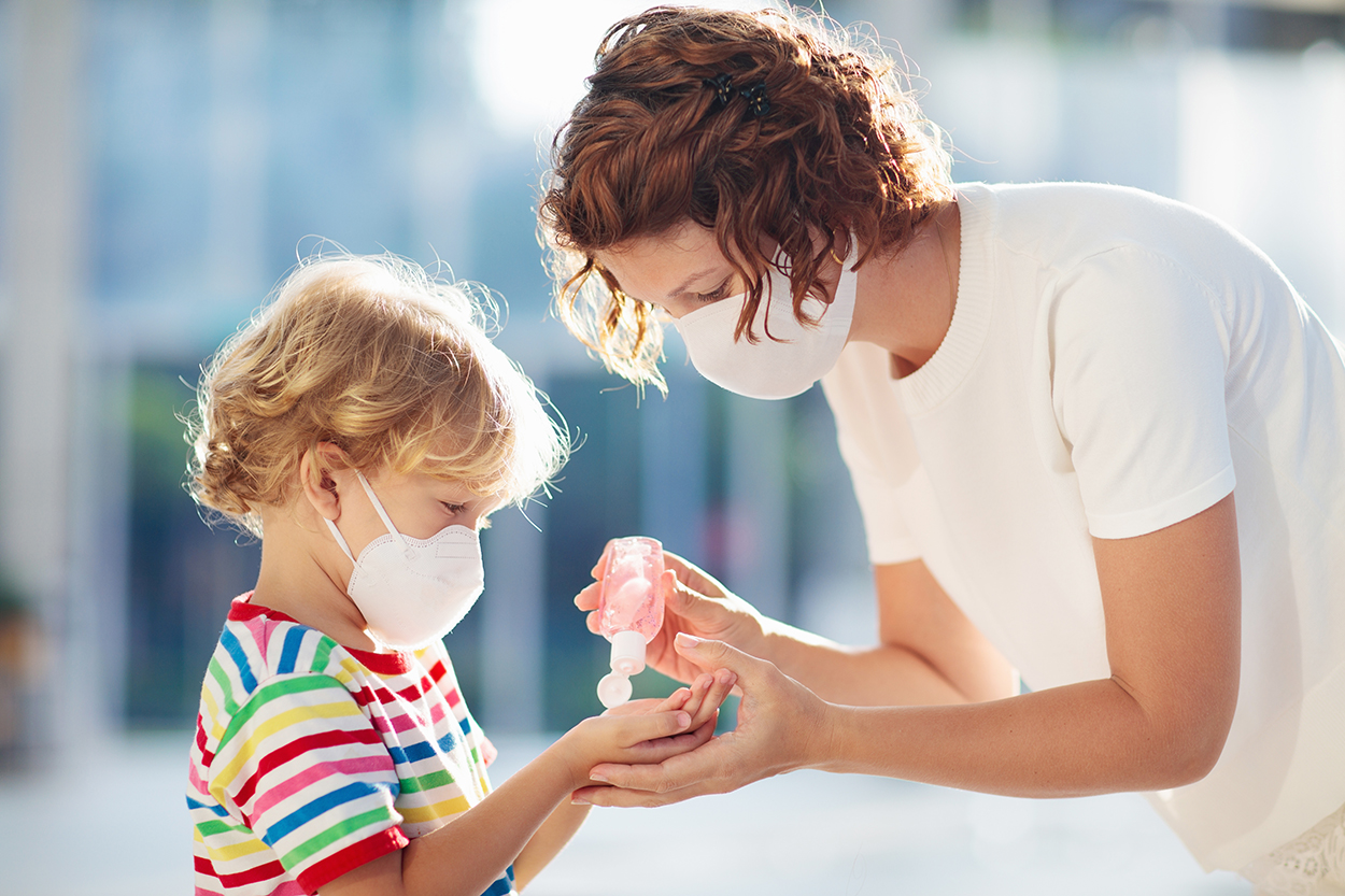 Child in mask receiving hand sanitizer from adult outdoors.