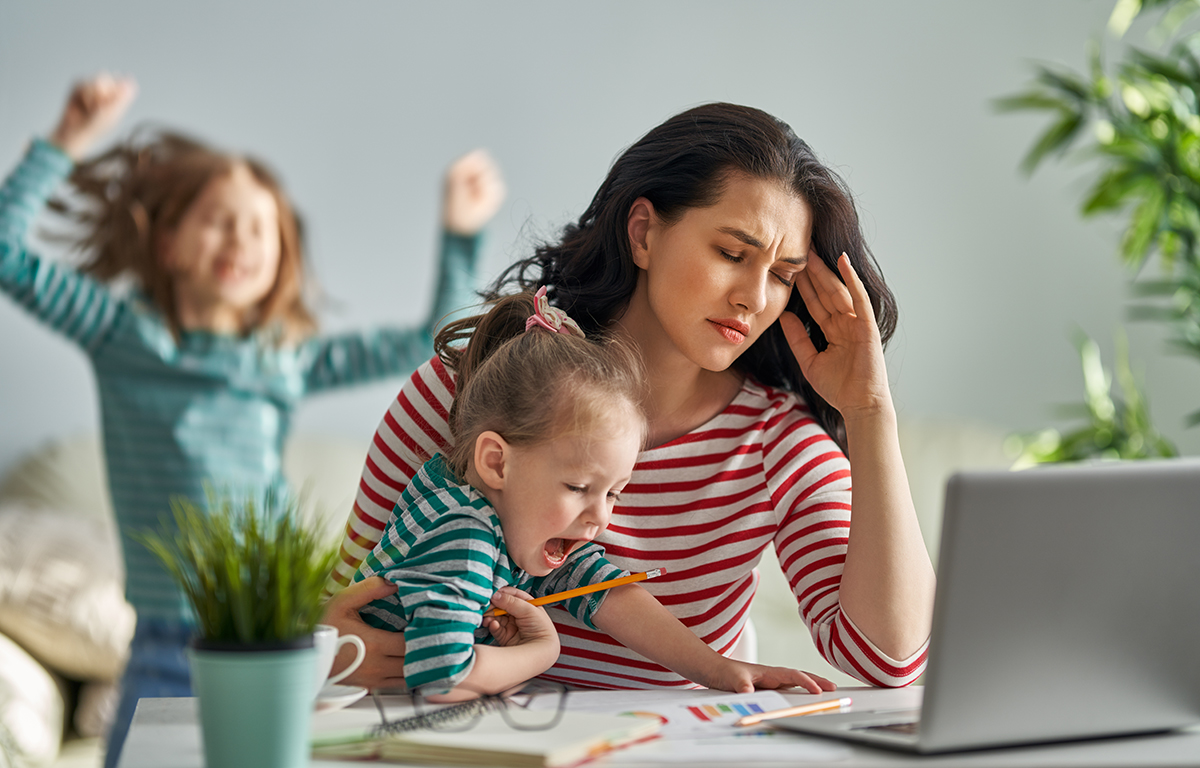 Stressed mother working on laptop with noisy children in background.