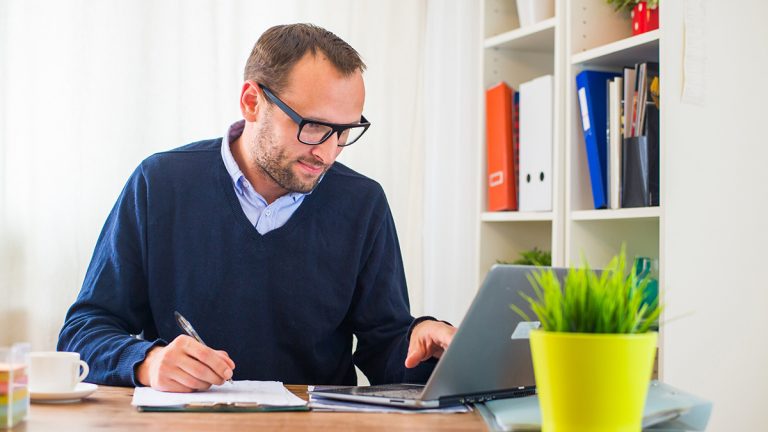 Focused man working from home, writing notes beside a laptop in a bright and organized home office.