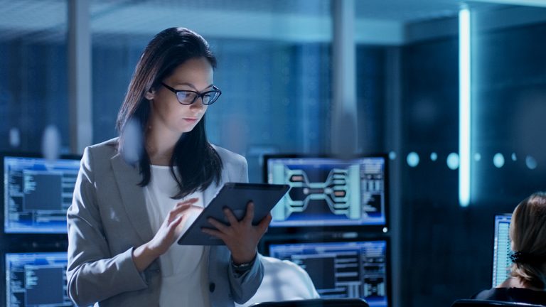 Professional woman using a tablet in a high-tech server room with digital screens in the background.