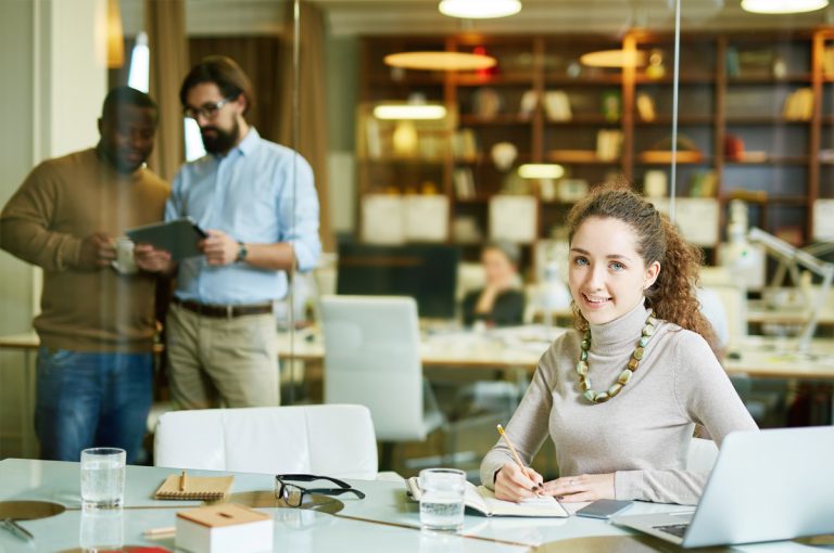 Young woman taking notes at her desk in a vibrant office, with two male colleagues discussing in the background.