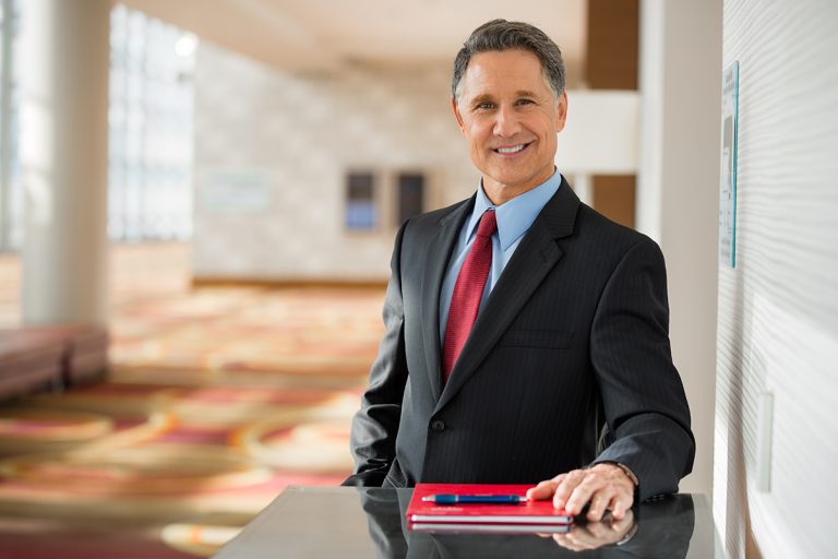 Confident businessman in a suit standing with a notebook and pen in a modern office lobby.