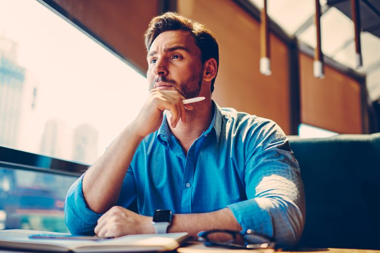 Thoughtful man in a blue shirt sitting by a window with a pen and notebook.