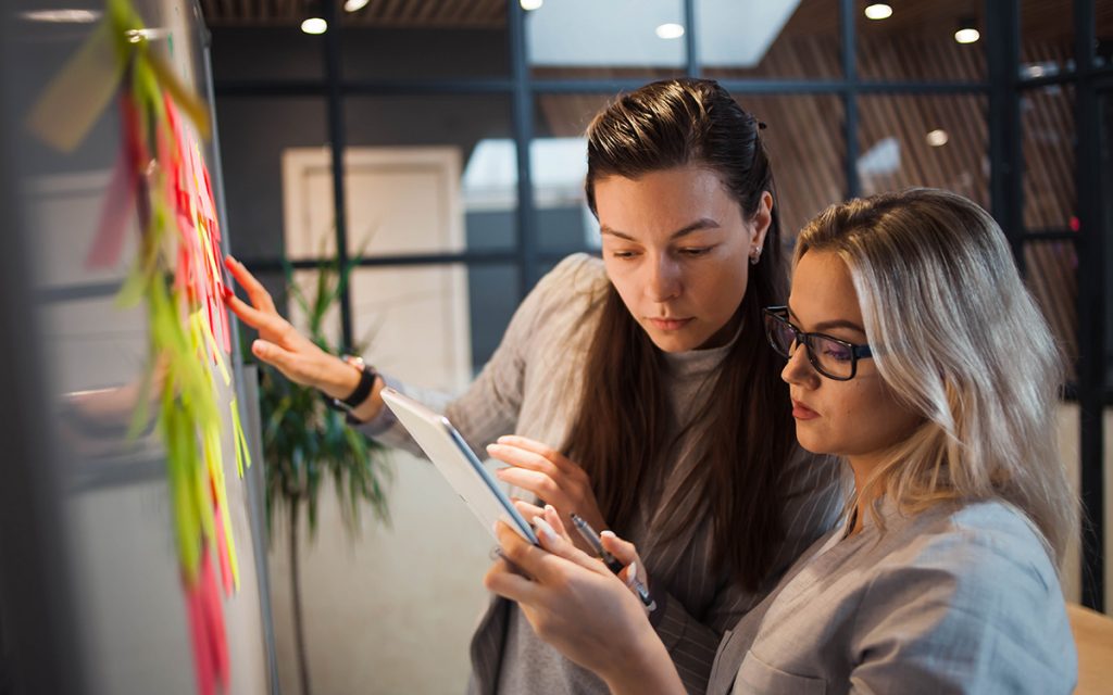 Two women collaborating and discussing ideas using a tablet in front of a board with sticky notes.