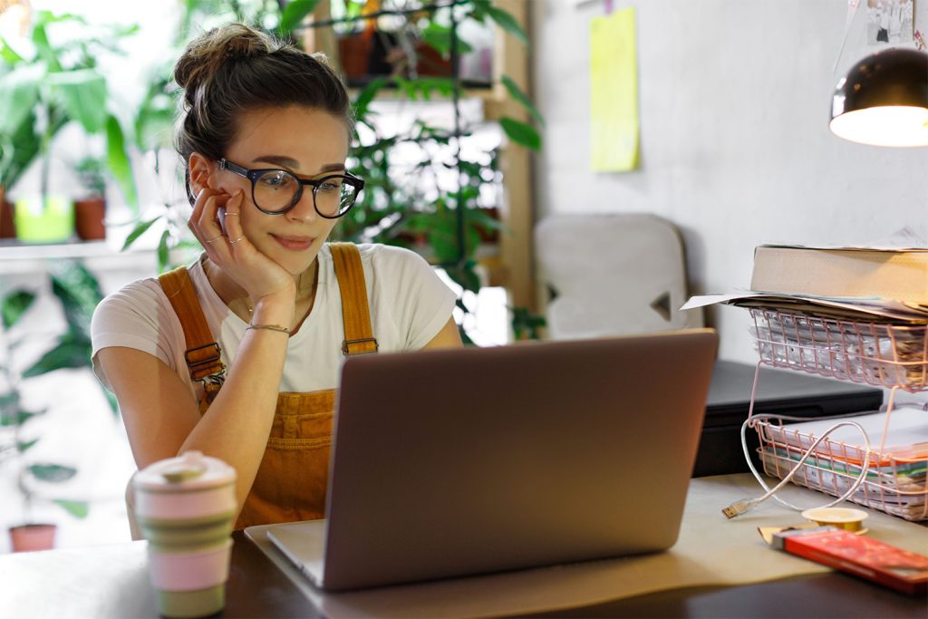 Young woman with glasses working on a laptop at home, surrounded by plants and office supplies.