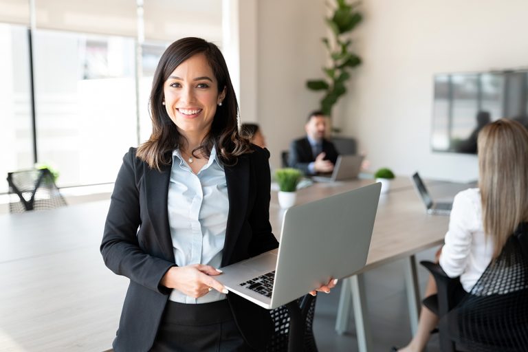 Professional woman with laptop smiling in office, colleagues in background.