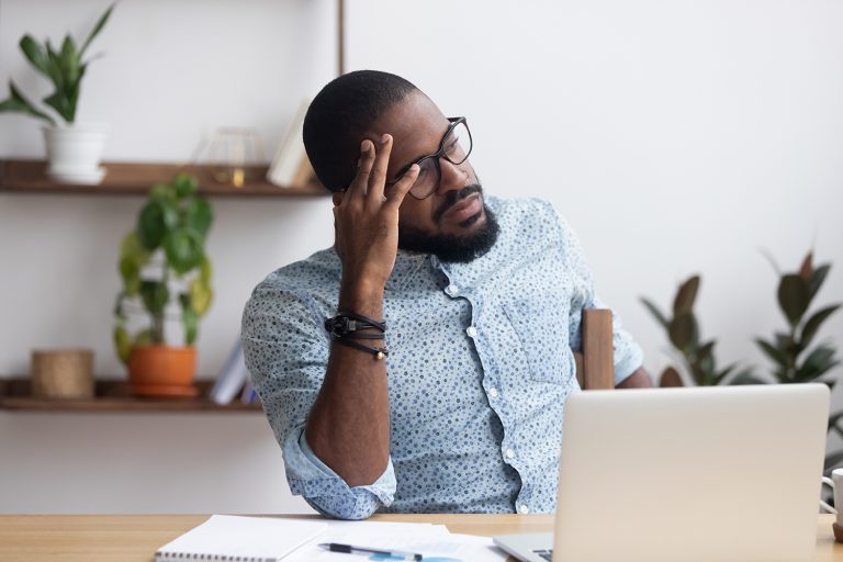 Pensive African American man with glasses looking at his laptop, sitting at a home office desk.