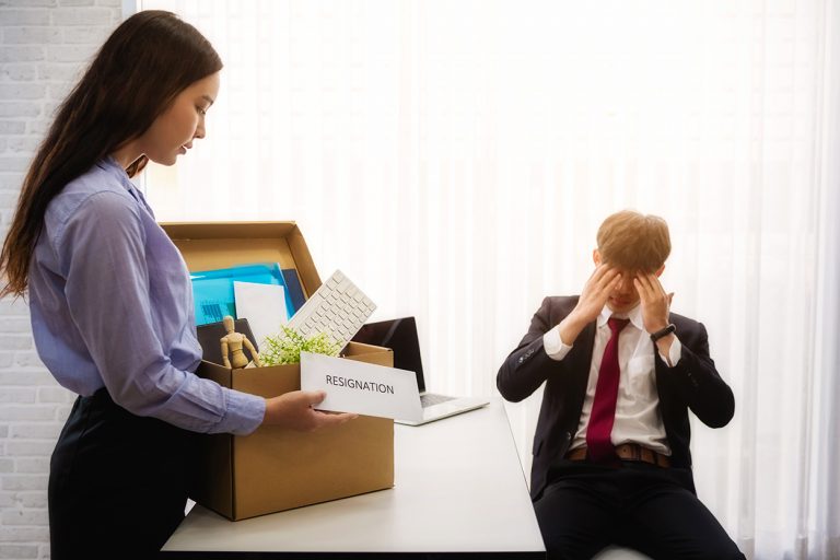 Woman holding a box with her belongings as she resigns, with a distraught male colleague in the background.