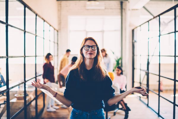 Welcoming young woman with glasses standing with arms outstretched in a busy modern office environment.