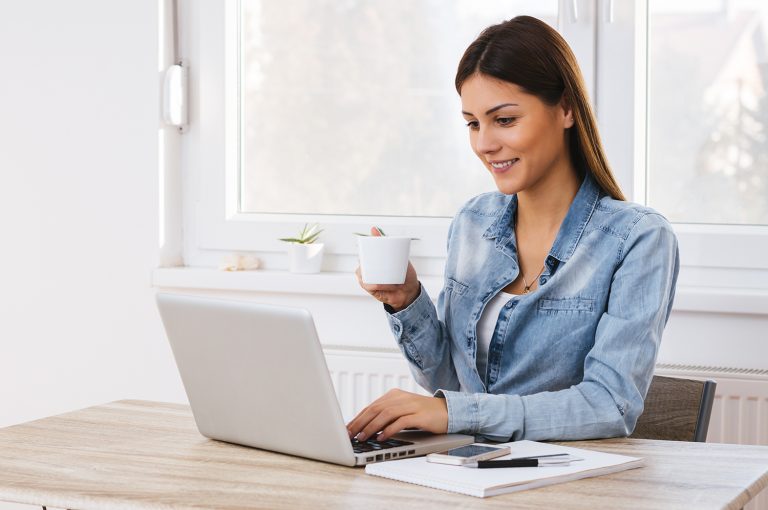 Smiling woman using a laptop while holding a coffee cup in a bright home office setting.