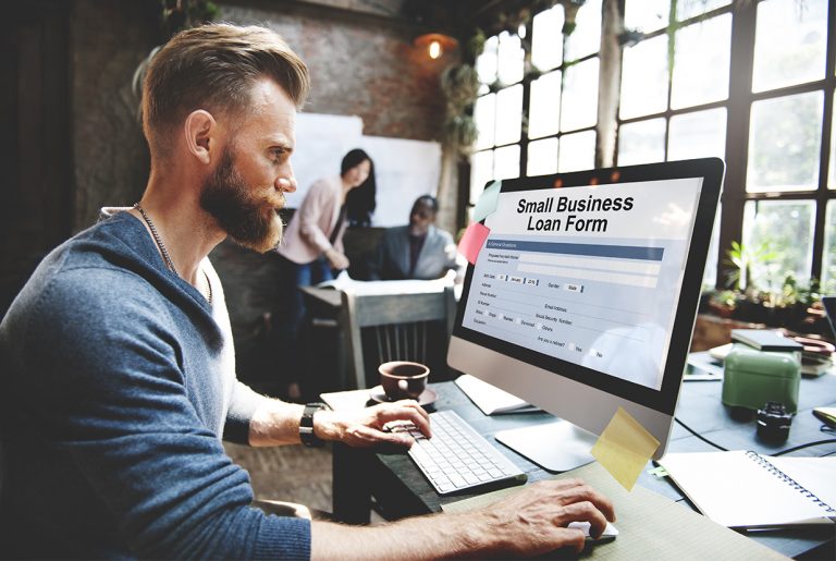 Bearded businessman applying for a small business loan online, with a team meeting in the background.