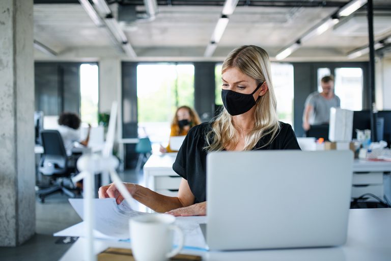 Focused woman with a face mask working at her laptop in a modern office with colleagues around.