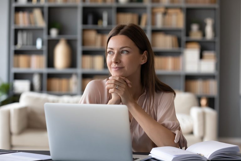 Pensive young woman looking thoughtful while working on a laptop in a home office with bookshelves.