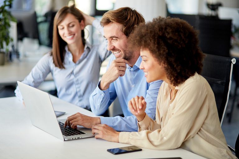 Three cheerful professionals working together on a laptop in a bright modern office setting.