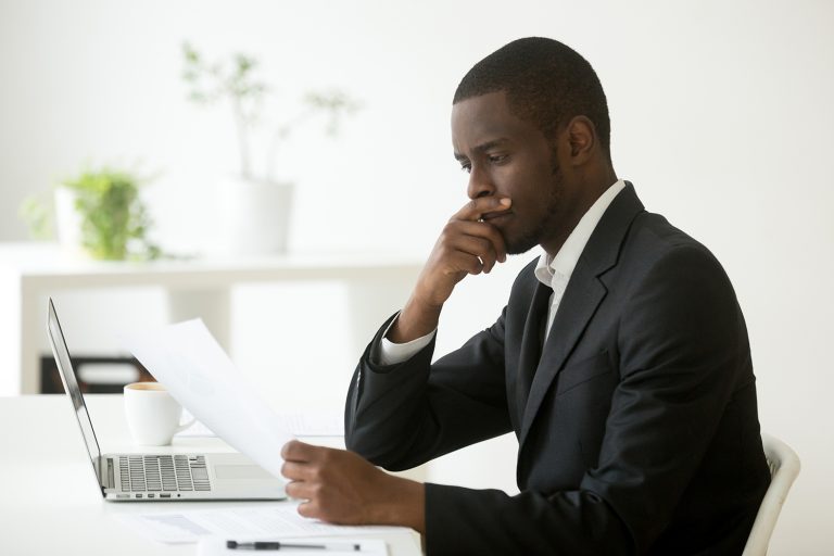 Serious African American businessman analyzing documents while working on a laptop in a modern office.