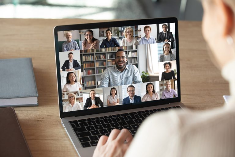 View over a woman's shoulder of a diverse virtual meeting with multiple colleagues on her laptop screen.
