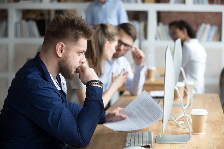 Focused man reading a document at his computer with colleagues discussing in the background at the office.