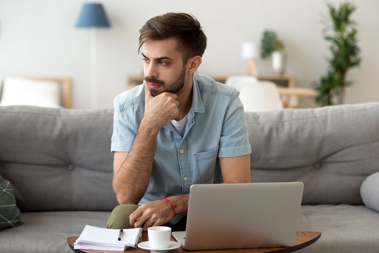 Focused young man with a beard working from a laptop on a couch in a cozy home setting.