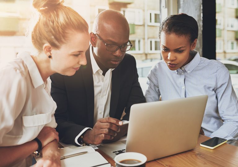 Diverse business team closely examining data on a laptop in a sunlit, informal office setting.