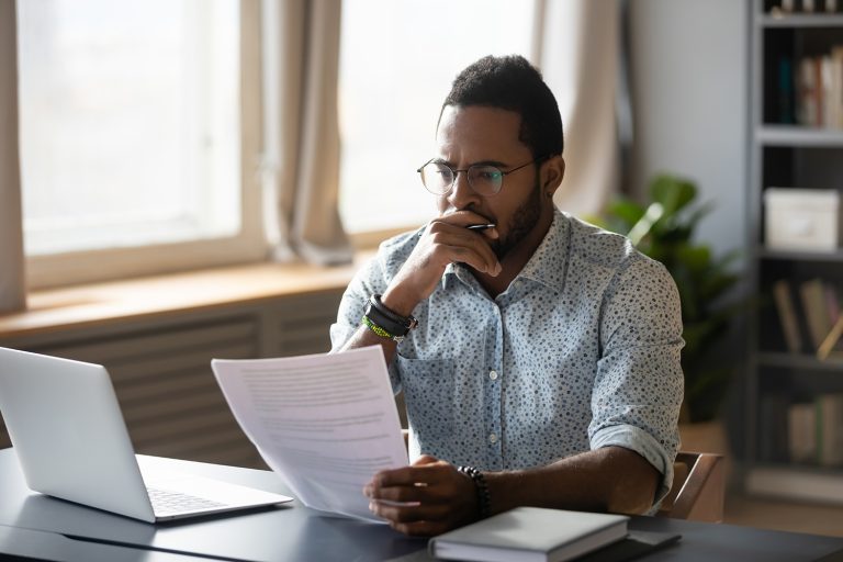 Concentrated African American man reviewing documents while working on a laptop in a well-lit office.