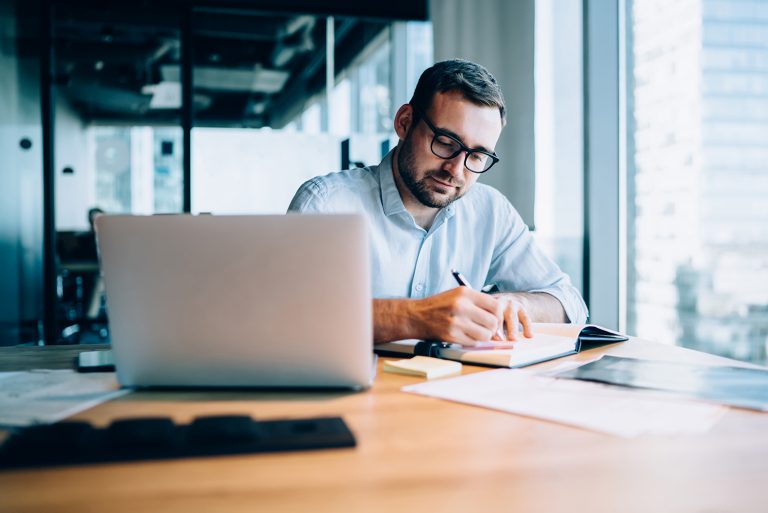 Focused businessman writing notes while working on a laptop in a modern office with city views.