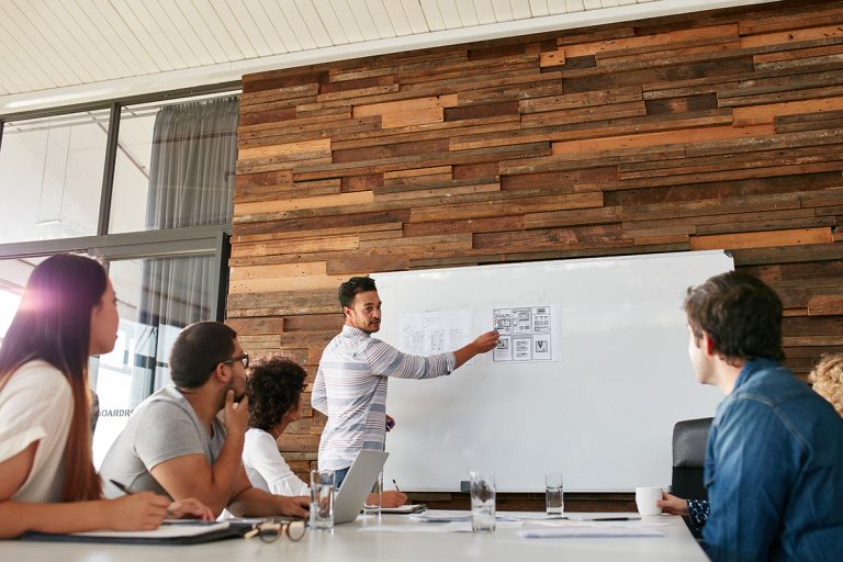 Diverse business team attentively listening to a male presenter at a whiteboard in a wooden-paneled meeting room.