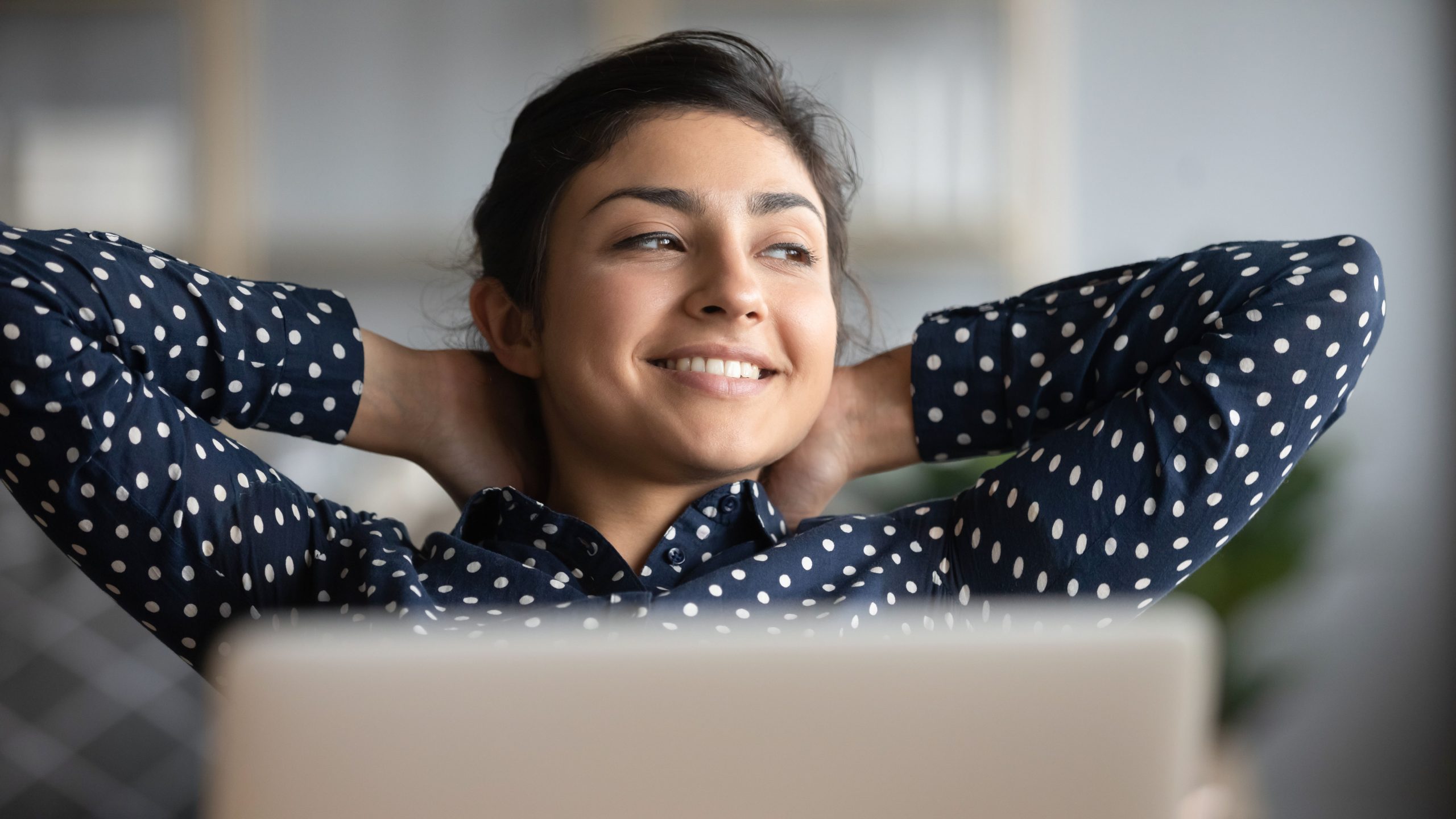 Content young woman in a polka-dot shirt relaxing with hands behind head, gazing away, in front of a laptop.