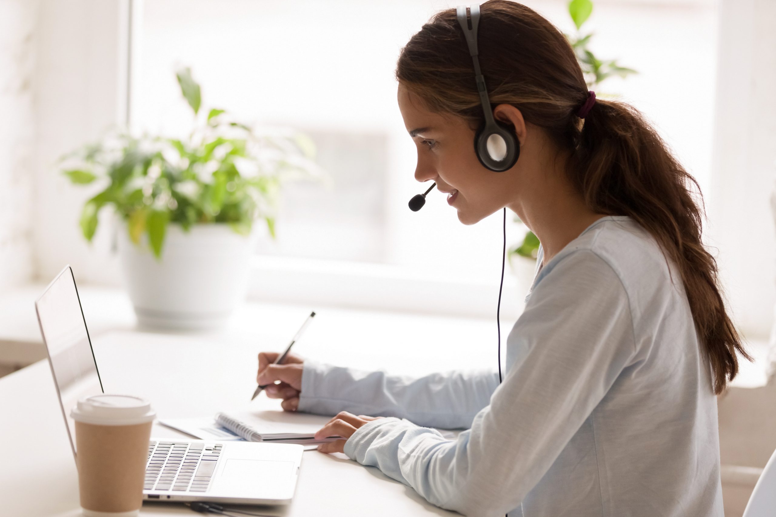 Focused woman with headset writing notes at laptop in bright office.